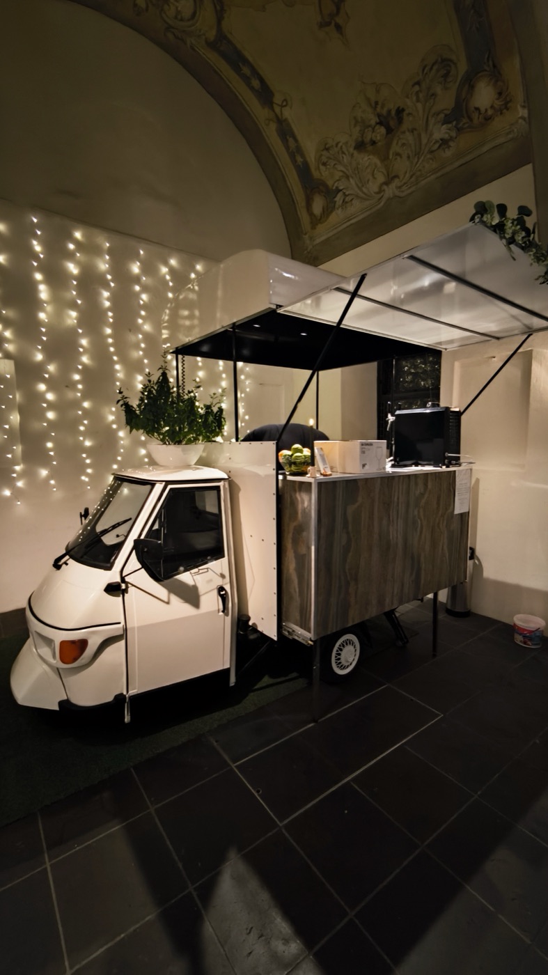 Die cremeweiße Piaggio Ape vor einem Vorhang aus Lichterketten