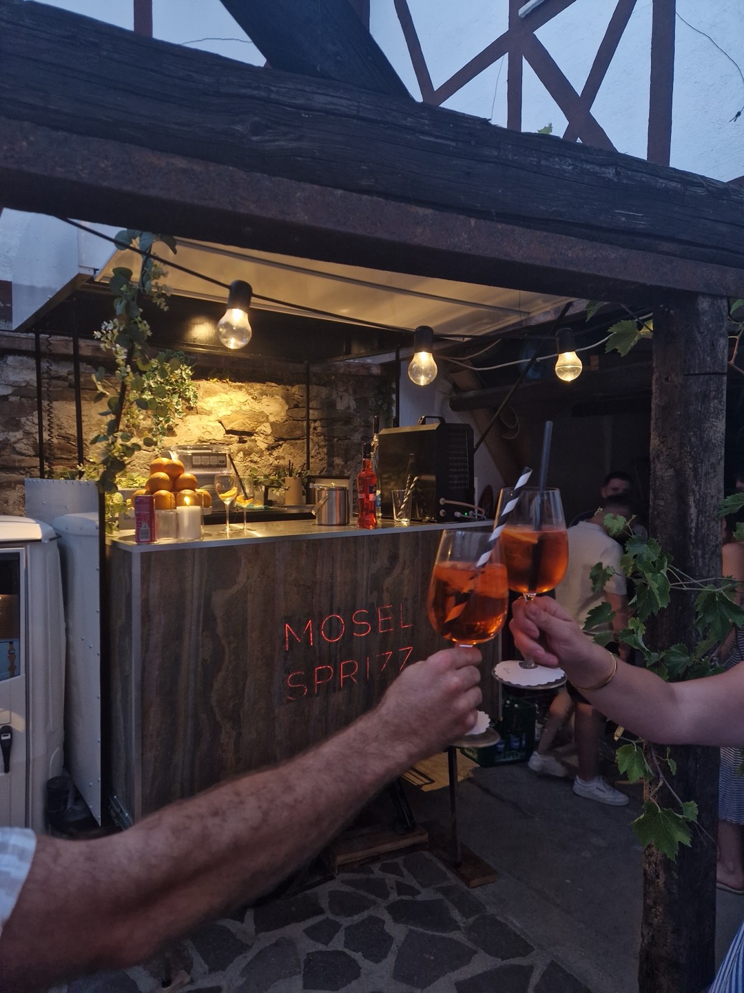 Zwei Gäste stoßen mit Aperol Spritz vor der Mosel-Sprizz-Bar mit rotem Leuchtschriftzug an