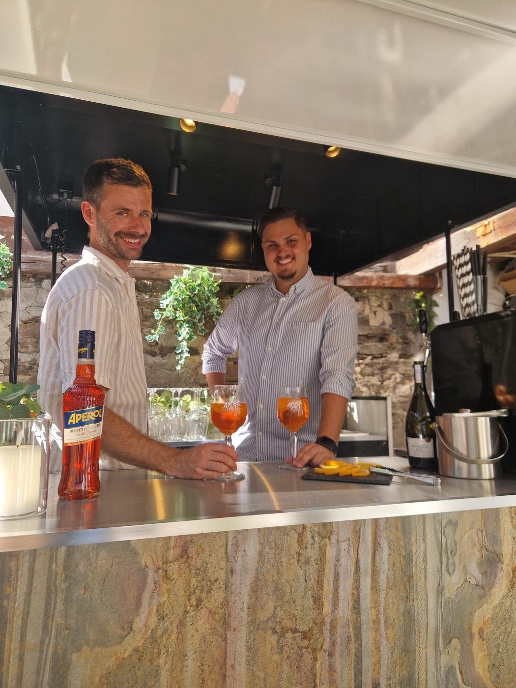 Die beiden Gründer von Mosel Sprizz lachen hinter der Bar der Ape, vor ihnen zwei Aperol Spritz